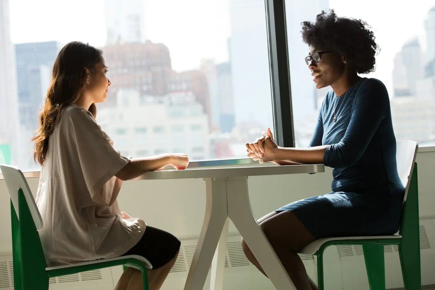 Two women discussing their work at a table beside a bright office window