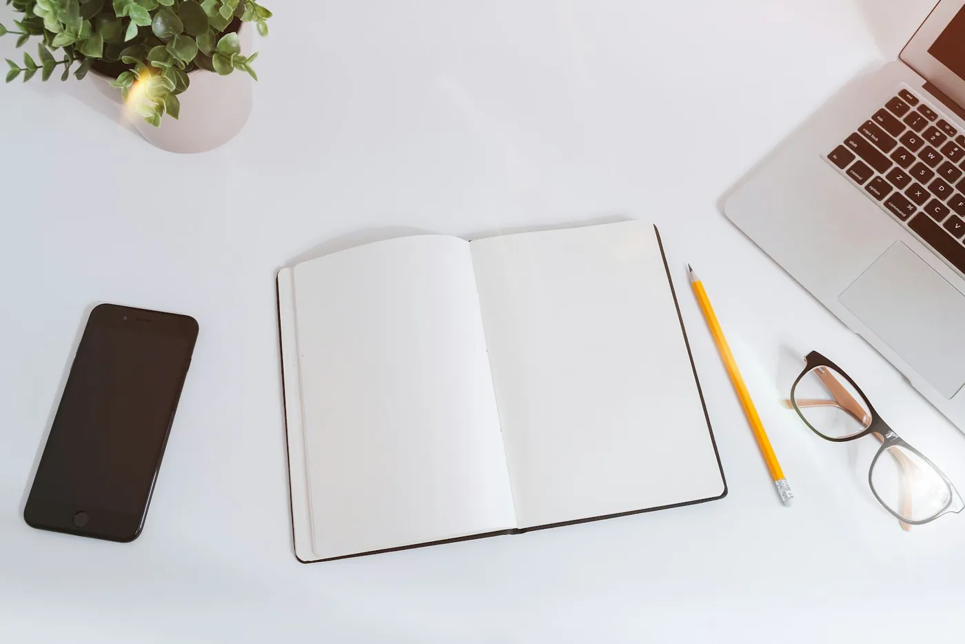 An open notebook, phone, laptop and glasses on a white desk